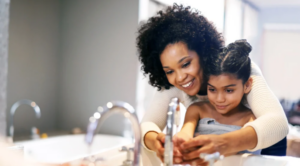 Mother helping young daughter wash her hands in a sink. View of them in mirror above sink.