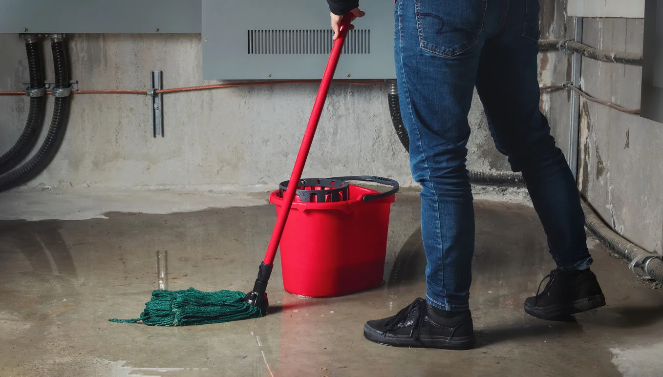 Person wearing jeans  and black sneakers, cleaning up a flooded concrete basement floor with a mop and red bucket.